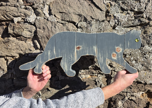 Stalking cat rustic wooden wall art being held with hands for sizing against a stone wall 