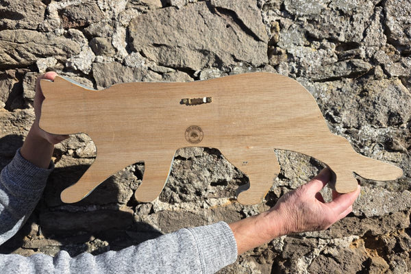 Reverse side of Stalking cat rustic wooden wall art being held with hands for sizing against a stone wall 