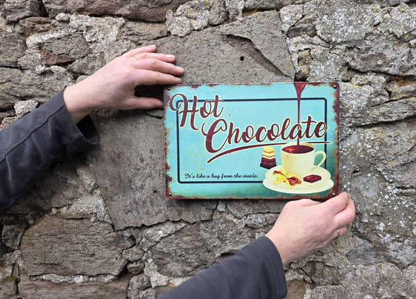 VINTAGE STYLE METAL SIGN (REPRODUCTION) HOT CHOCOLATE HANGING ON A STONE WALL SHOWING SIZING OF THE SIGN BY AN ADULT