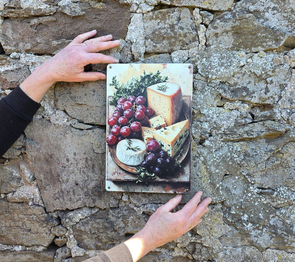 A decorative vintage style metal sign (repro) hanging on a stone wall with an image of cheeses and grapes on a cheeseboard, being held to show sizing of the sign