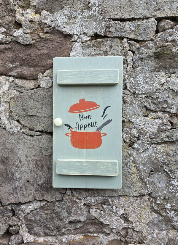 Vintage style shabby chic, sage green pine wall art shutter hanging on an old stone wall, with a design on it of a red crock-pot & lid, along with the words "Bon Appetit" in black text