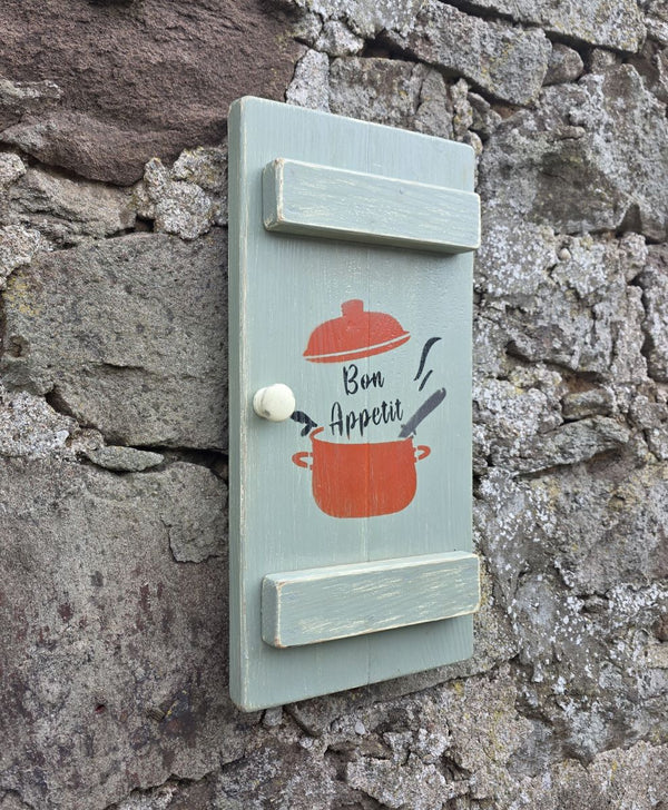 Side angle view of a Vintage style shabby chic, sage green pine wall art shutter hanging on an old stone wall, with a design on it of a red crock-pot & lid, along with the words "Bon Appetit" in black text