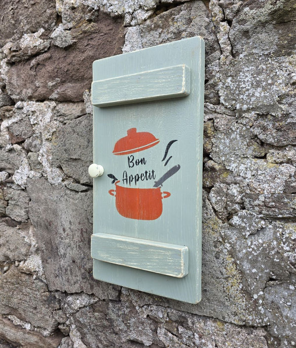 Another side angle view of a Vintage style shabby chic, sage green pine wall art shutter hanging on an old stone wall, with a design on it of a red crock-pot & lid, along with the words "Bon Appetit" in black text