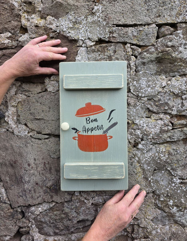 a Vintage style shabby chic, sage green pine wall art shutter being held up against an old stone wall, with a design on it of a red crock-pot & lid, along with the words "Bon Appetit" in black text
