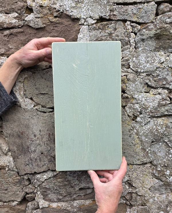 The reverse-side of a Vintage style shabby chic, sage green pine wall art shutter being held up against an old stone wall