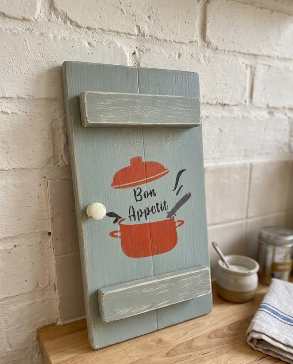 a Vintage style shabby chic, sage green pine wall art shutter with a design on it of a red crock-pot & lid, along with the words "Bon Appetit" in black text. The shutter is leaning against a rustic style kitchen wall & standing on top of a wooden worktop