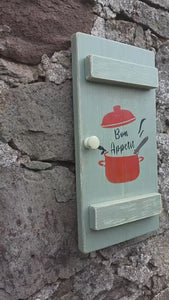 a Vintage style shabby chic, sage green pine wall art shutter hanging on an old stone wall, with a design on the shutter of a red crock-pot & lid, along with the words "Bon Appetit" in black text